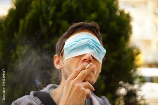 Fototapeta A portrait of a man with a face mask on his eyes. He is smoking a cigarette.