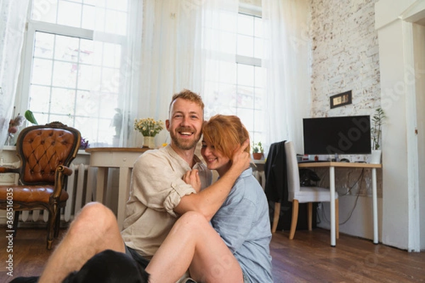 Fototapeta young couple sitting on the floor of their flat and holding each other. redhead woman and man smiling and spending leisure time together. laughing and happiness