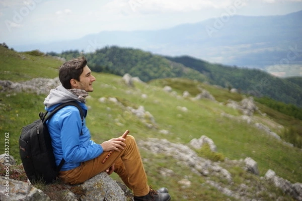 Fototapeta A young male tourist sitting on a rock. He is on the summit of Stara Planina (Balkan Mountains) and enjoying the view. In one of his hands, he's holding a wooden flute/ pipe.