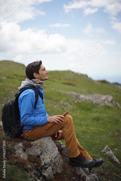 Fototapeta A young male tourist sitting on a rock. He is on the summit of Stara Planina (Balkan Mountains) and enjoying the view. In one of his hands, he's holding a wooden flute/ pipe.