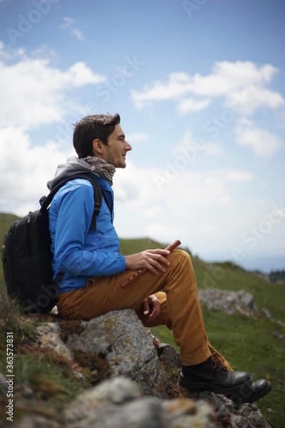 Fototapeta A young male tourist sitting on a rock. He is on the summit of Stara Planina (Balkan Mountains) and enjoying the view. In one of his hands, he's holding a wooden flute/ pipe.