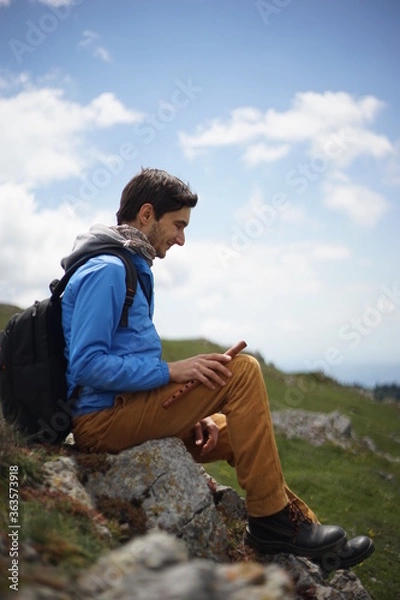 Fototapeta A young male tourist sitting on a rock. He is on the summit of Stara Planina (Balkan Mountains) and enjoying the view. In one of his hands, he's holding a wooden flute/ pipe.