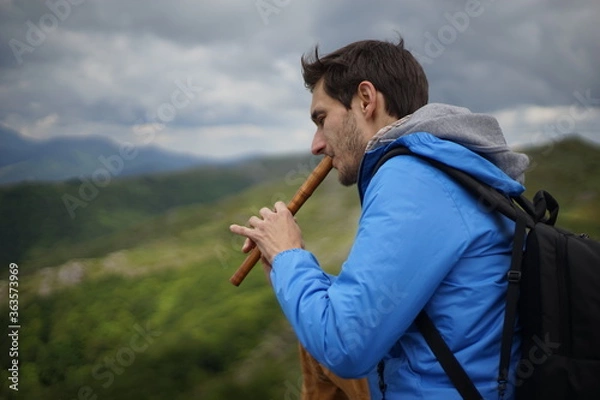 Fototapeta A side view of a young male tourist playing a wooden reed-pipe/ flute. He is on the summit of Stara Planina (Balkan Mountains). 