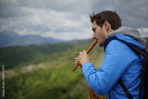 Fototapeta A side view of a young male tourist playing a wooden reed-pipe/ flute. He is on the summit of Stara Planina (Balkan Mountains). 