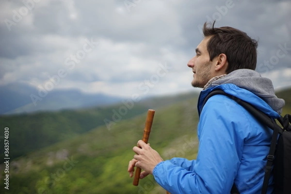 Fototapeta A side view of a young male tourist playing a wooden reed-pipe/ flute. He is on the summit of Stara Planina (Balkan Mountains). 