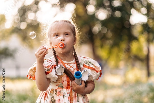 Fototapeta A girl with Down syndrome blows bubbles. The daily life of a child with disabilities. Chromosomal genetic disorder in a child.