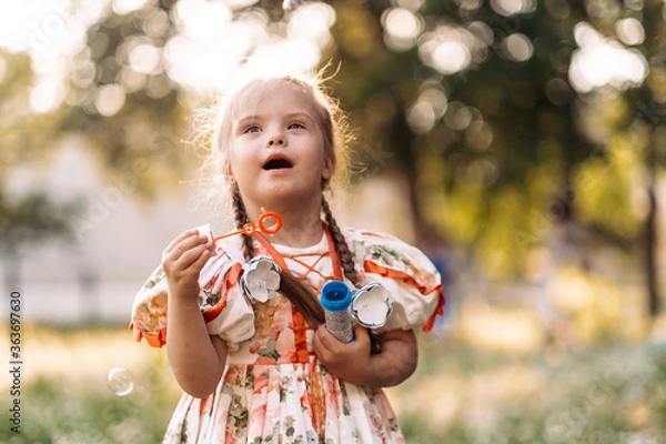 Fototapeta A girl with Down syndrome blows bubbles. The daily life of a child with disabilities. Chromosomal genetic disorder in a child.