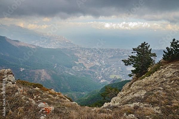 Fototapeta View from the mountain edge to the Yalta city in Wuchang-Su Valley on the Black Sea coast in south Crimea. Dry grass and stones in the foreground. Forest on the slope. City on the shore. Cloudy sky.