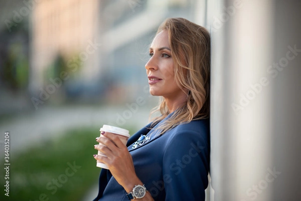 Fototapeta Portrait of a middle-aged woman with a disposable glass with a white plastic cup in her hand in an expensive watch, standing alone in the evening light against the background of houses in the city.
