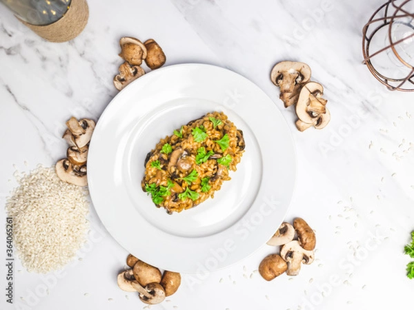Fototapeta Mushroom risotto with Parmesan cheese and white Wine in a white serving plate on a white marble table with risotto Rice and raw mushroom on the side. Free space for your text/decoration.