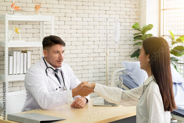 Obraz Caucasian male doctor with a stethoscope giving a handshake to his patient in white tone clinic.