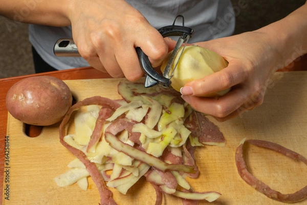 Obraz peeling potatoes with a knife for peeling vegetables. female hands peel potatoes over a wooden kitchen board while cooking