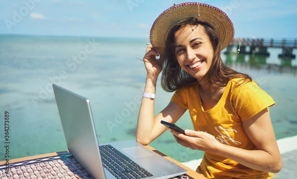 Obraz portret szczęśliwa freelancer kobieta w kapeluszu uśmiecha się do kamery, trzymając smartfon, pracując na komputerze przenośnym nad brzegiem morza. Wolność, praca zdalna, praca na własny rachunek, podróże i wakacje