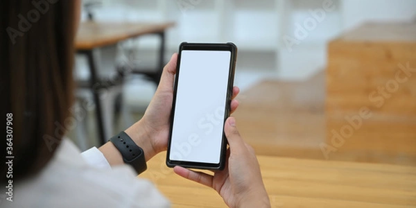 Fototapeta Cropped image hands are holding a white blank screen smartphone in the comfortable living room.