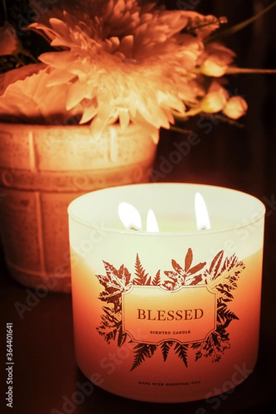 Fototapeta Lit scented candle with "Blessed" text, pot of flowers in the background
