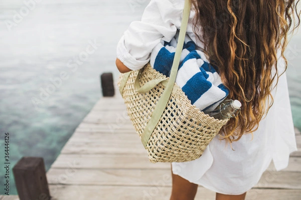 Fototapeta Woman with handmade wicker bag, two beach towels and glass bottle for water going to the beach. View from behind close up. Eco friendly and zero waste concept.