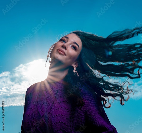 Obraz Happy Woman With Curly Fluttering in The Wind Hair at Blue Bright Sky Background. Summer Cotton Knitted Violet Sweater or Jumper