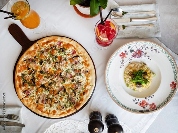 Obraz Dinner at an Italian restaurant. White tablecloth, on a pitcher’s table with ham and mushrooms and risotto with sea products. Two cold lemonade. Italian traditional food.