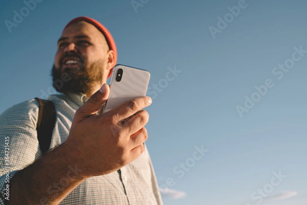 Fototapeta Caucasian male tourist holding modern cellphone gadget in hand and looking around during sunny day for recreation on nature, millennial blogger using digital technology for online web mobility