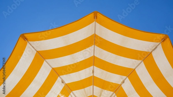 Fototapeta White and yellow striped beach umbrella. Blue sky in the background. View from below. Relaxing context. Summer holidays by the sea. General contest and location