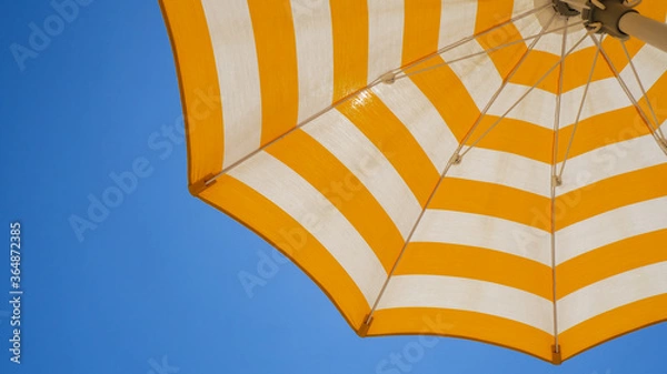 Fototapeta White and yellow striped beach umbrella. Blue sky in the background. View from below. Relaxing context. Summer holidays by the sea. General contest and location