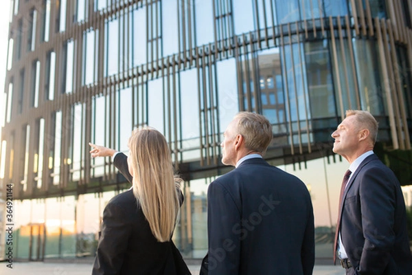 Fototapeta Businesspeople in suits pointing and looking at office building, discussing real property. Back view, copy space. Commercial real estate concept