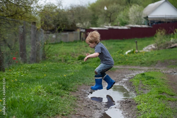 Fototapeta a small boy in rubber boots jumps through a muddy puddle