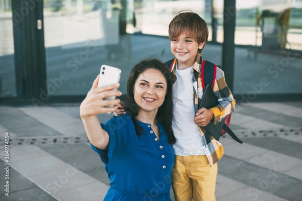 Fototapeta Mom and child with a backpack take selfie on the phone before going to school or kindergarten. Happy parents and children. Back to school. Side view