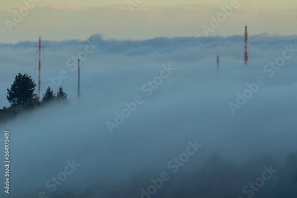 Fototapeta The fog covers a forest covering the trees and the telecommunication antennas on the hill