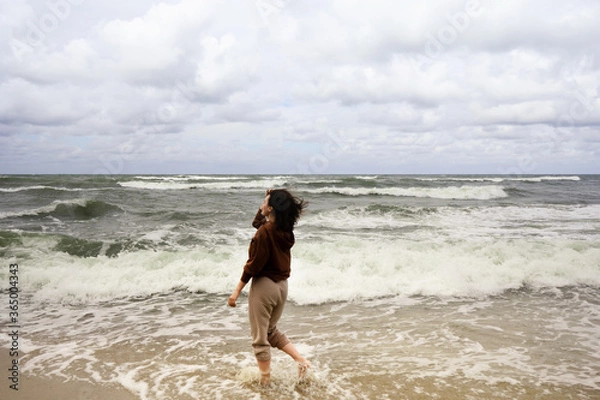 Fototapeta young woman walking on the beach with her feet in the water, watching tide on a Baltic Sea, windy day with waves, day on a shore, enjoying nature, relaxing outside by seaside 