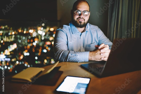 Fototapeta Portrait of prosperous businessman sitting at desktop in apartments with night city window view and modern technology, male freelancer looking at camera staying late for distance job online