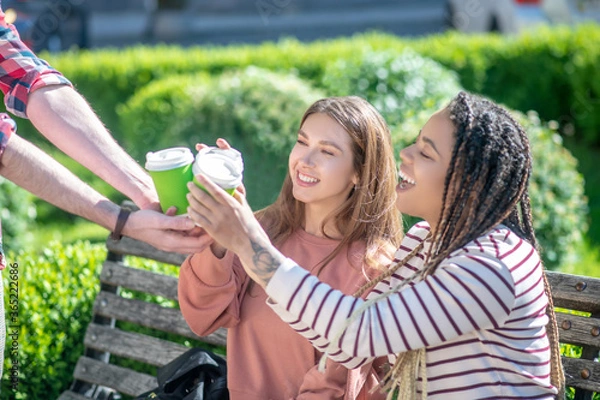 Fototapeta Two girls on bench taking coffee from hands of guy