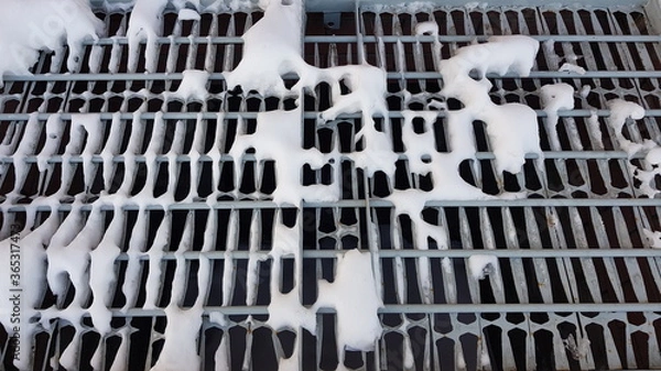 Fototapeta Close-up of white pieces of snow on a metal grate. Snow falls through the grating, revealing voids. Abstraction of snow and metal lattice. On a winter day, outdoors in the evening. Texture.