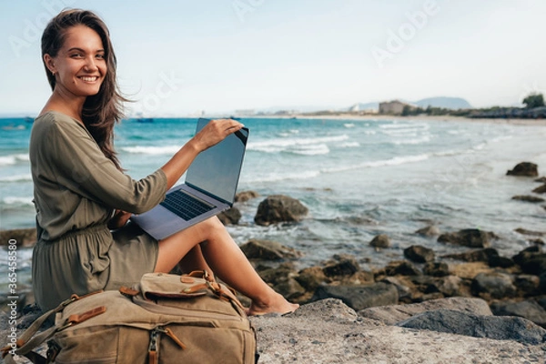 Obraz Young female freelancer working on a laptop while traveling by the sea, remote work concept. Work by the sea. Using modern technology, the Internet for communication