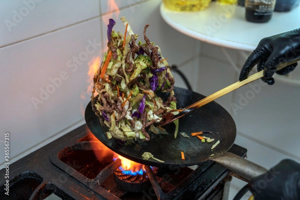 Obraz Chef making japonese food. Stir-fry soba noodles with beef and vegetables in wok pan on dark background