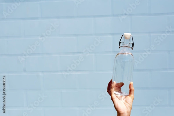 Fototapeta A woman holds a clear glass water bottle in her hand for personal use. On a bright sunny day. Healthy lifestyle and eco friendly reusable and recycle concept. Light blue brick wall background.