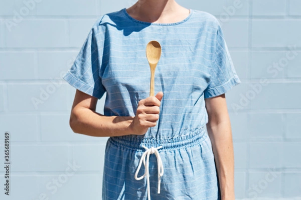 Fototapeta A woman holds a wooden bamboo spoon in her hand in front of her, ready to eat. On a bright sunny day. Healthy lifestyle and eco friendly, reusable and recycle concept. Blue brick wall behind.