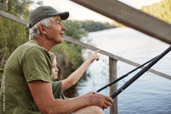 Fototapeta Senior man with his grandson sitting on wooden pontoon with fishing rods in hands, enjoying beautiful nature, little boy pointing at something with his finger.