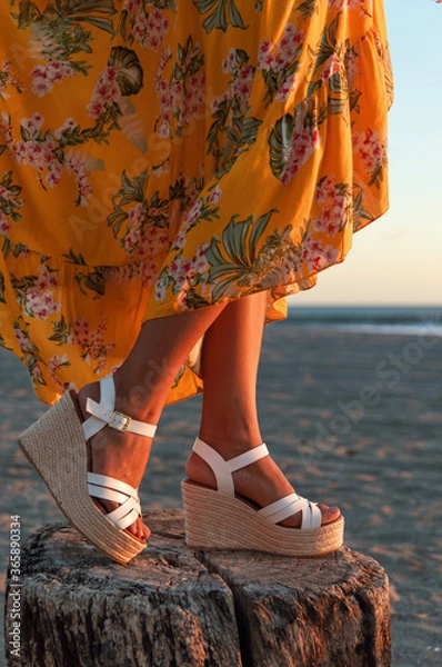 Obraz Legs of a female in a yellow dress with floral patterns and elegant white wedges at the beach