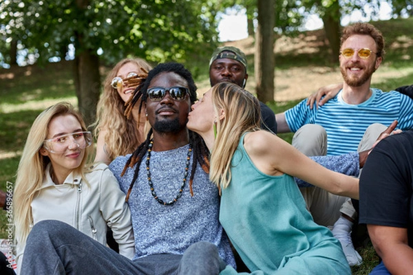 Fototapeta caucasian girl kisses black man in the park, group of friendly diverse people sit on the grass. make love, not war. harmonious relationships of american multiethnic people