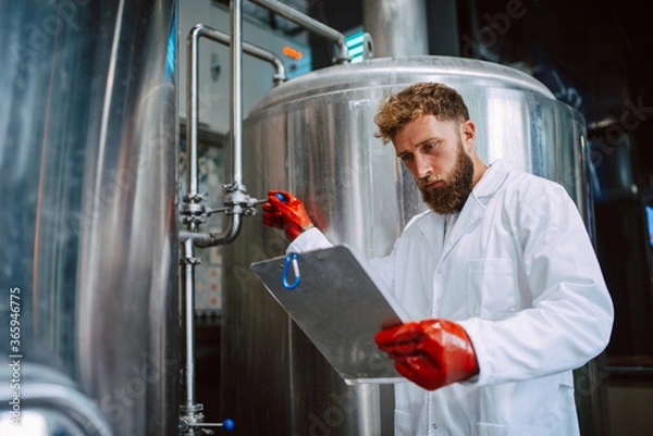 Fototapeta Caucasian male professional  technologist expert in white suit  operating production machine in food factory and doing quality control and holding checklist. Checking production in food industry.