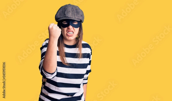 Fototapeta Young beautiful brunette woman wearing burglar mask angry and mad raising fist frustrated and furious while shouting with anger. rage and aggressive concept.