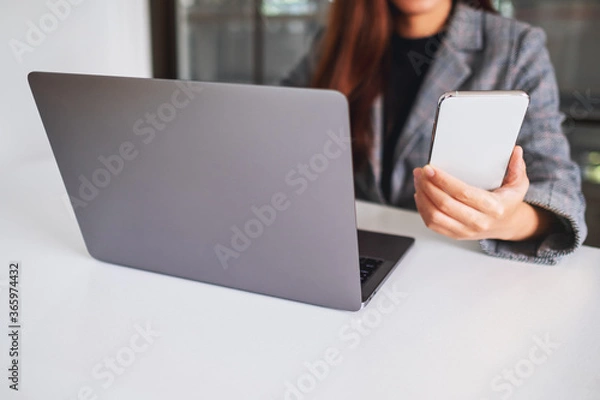 Fototapeta A young business woman holding and using mobile phone with laptop computer on the table in office