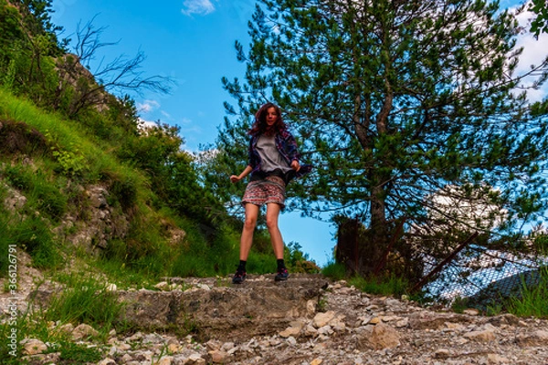 Fototapeta A full body shot of an energetic young Caucasian redhead woman jumping on a hiking path in the mountains under warm summer sunlight (Puget-Theniers, France)