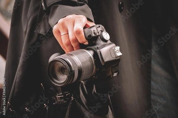 Obraz A girl photographer in a black raincoat in rainy weather holds a black umbrella and a SLR camera in her hand.