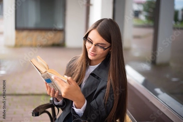 Obraz Beautiful woman in a eyeglasses and black coat is sitting outdoor and reading an interesting book.