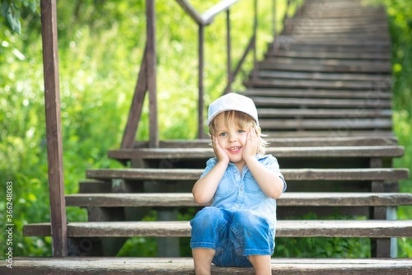 Obraz A boy in a white cap sits in a Park on a wooden staircase, posing and smiling.