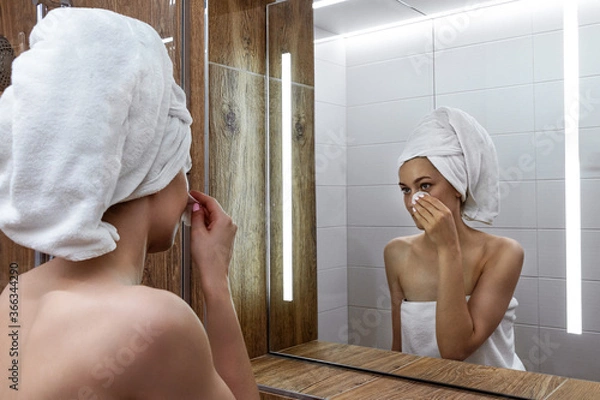 Obraz Young woman after a shower standing in the bathroom in front of the mirror with a towel wrapped on her head and washes her face with a cosmetic cotton pad with lotion. Skin care