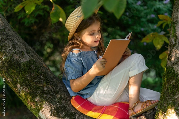 Obraz A cute little girl in a straw hat reading a book on a large tree. Children and reading. Concept of children's recreation without gadgets.