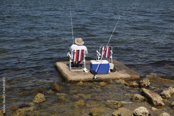 Fototapeta fisherman in a hat sitting on a chair with a red and white stripe is fishing with a fishing rod on the sea or lake there is a container for fish nearby. man sitting with his back.